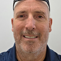 A man wearing a dark blue jacket over a black shirt, with sunglasses pushed up on his head, smiles at the camera. 