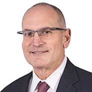 A man in a suit, wearing glasses, looks at the camera.