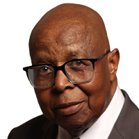 An older, bald man with brown skin wears a brown suit with a tie and glasses looks out at the camera. 