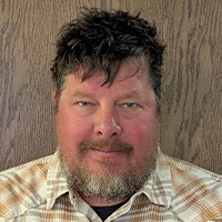 A man with spiky dark hair and a plaid shirt looks at the camera. 