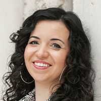 A woman with curly black hair, wearing a black jacket and hoop earrings, smiles at the camera. 