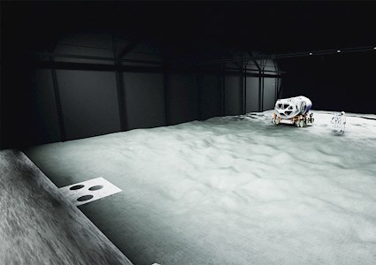 machine on wheels sitting on material that resembles the moon's surface  in a large dark hall that resembles