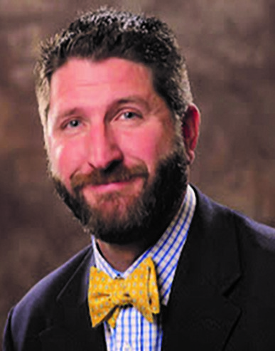 white man with brown beard and hair wearing a blue plaid shirt, a blue jacket and a yellow bow tie