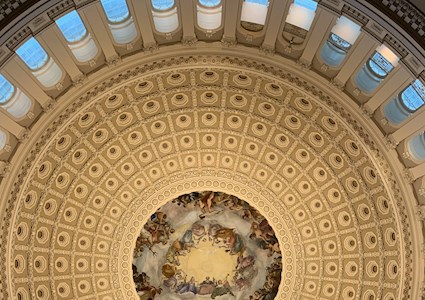 looking upward at the interior of the US Capitol dome's painting, surrounded by arched windows showing blue sky
