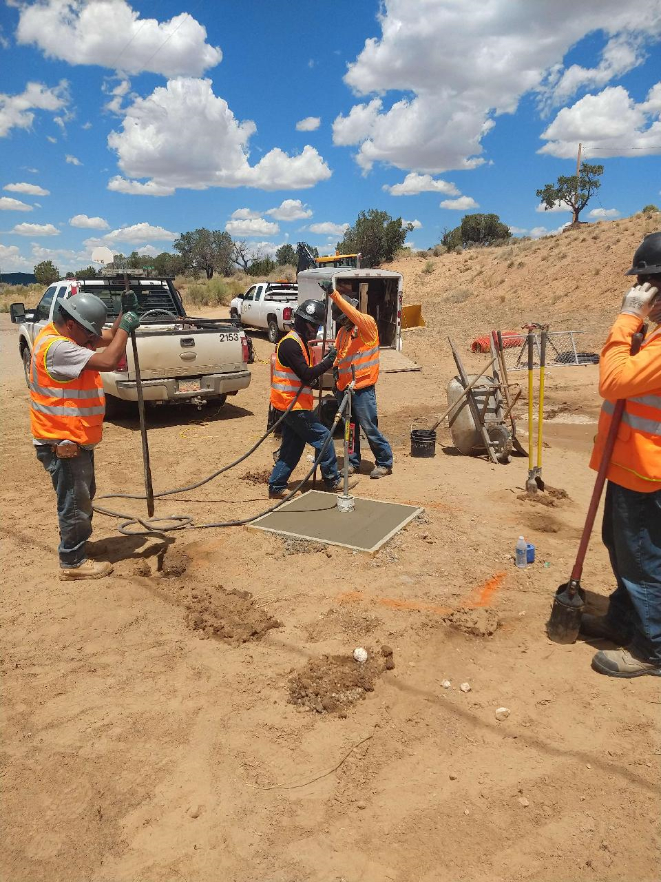 workers in orange vests stand in a sandy area while a square of concrete is placed