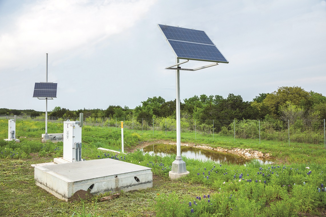photo of a solar panel placed on a pole in the ground that's near a small body of water like a pond