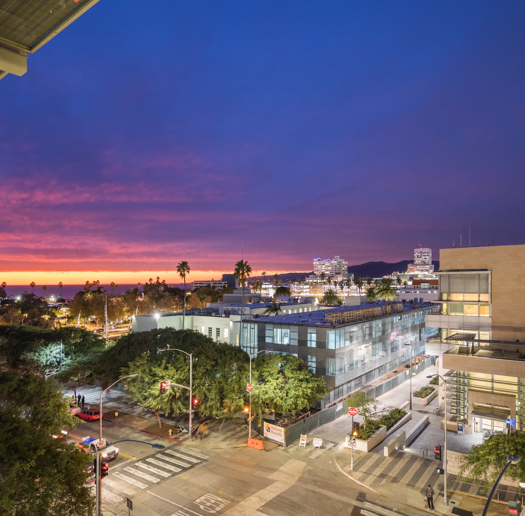 a glass building stands on a brightly lit street corner while a yellow to purple to deep blue sunset occurs beyond it