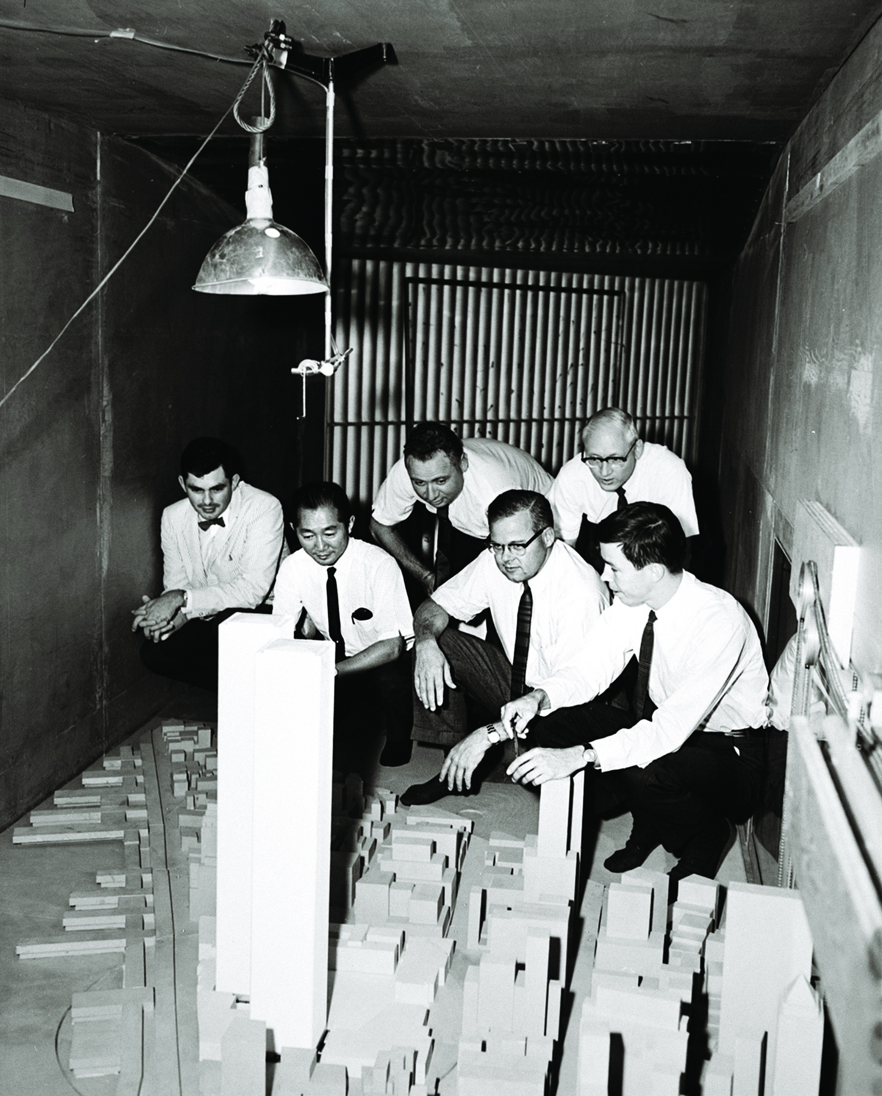 six men in white shirts and black ties hovering over a model of a large building complex