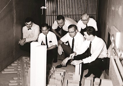 five men in white shirts and black ties hovering over a model of a large building complex