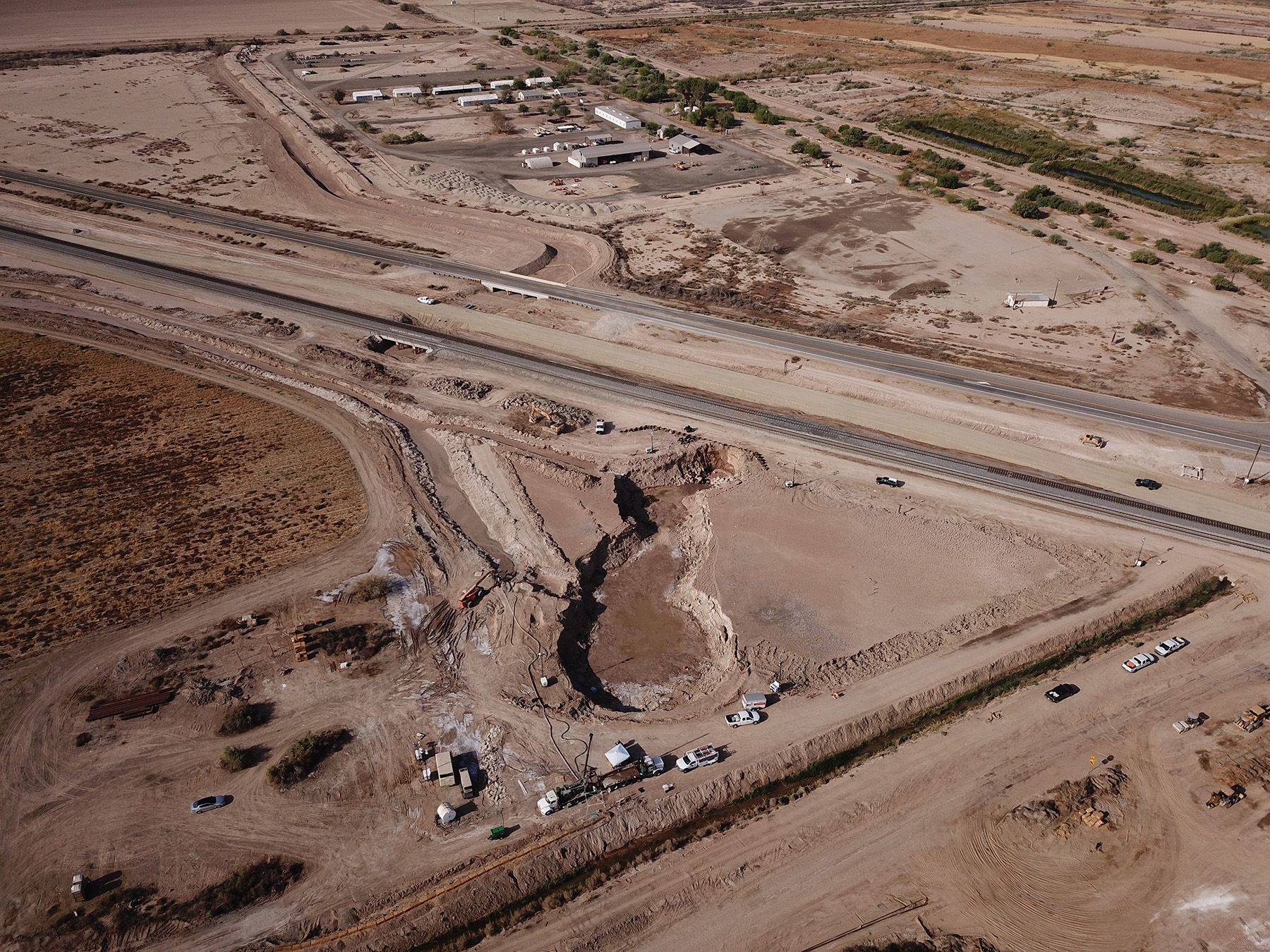 large dirt field with a two lane roadway dividing it. in the center foreground is a large irregular shaped depression filled with muddy water