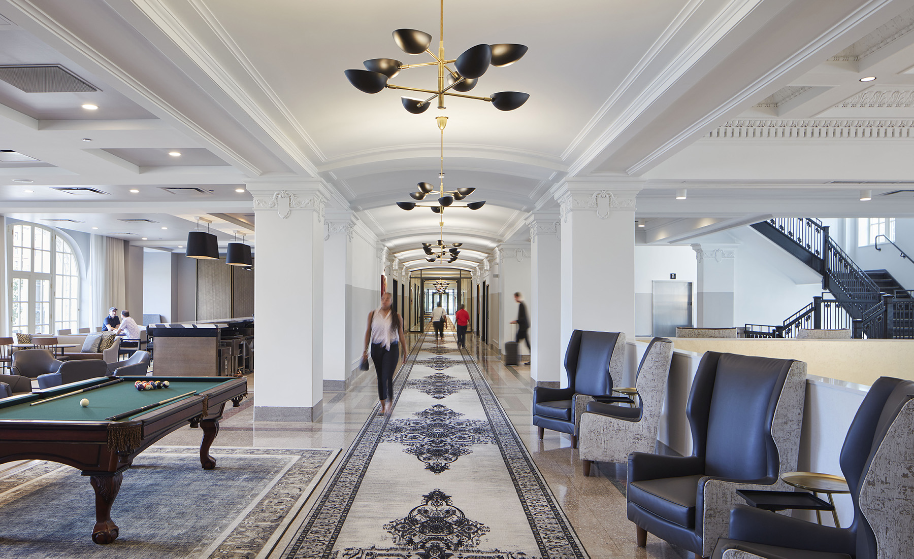 people walking down a hallway with marble floor, chairs, and carpet