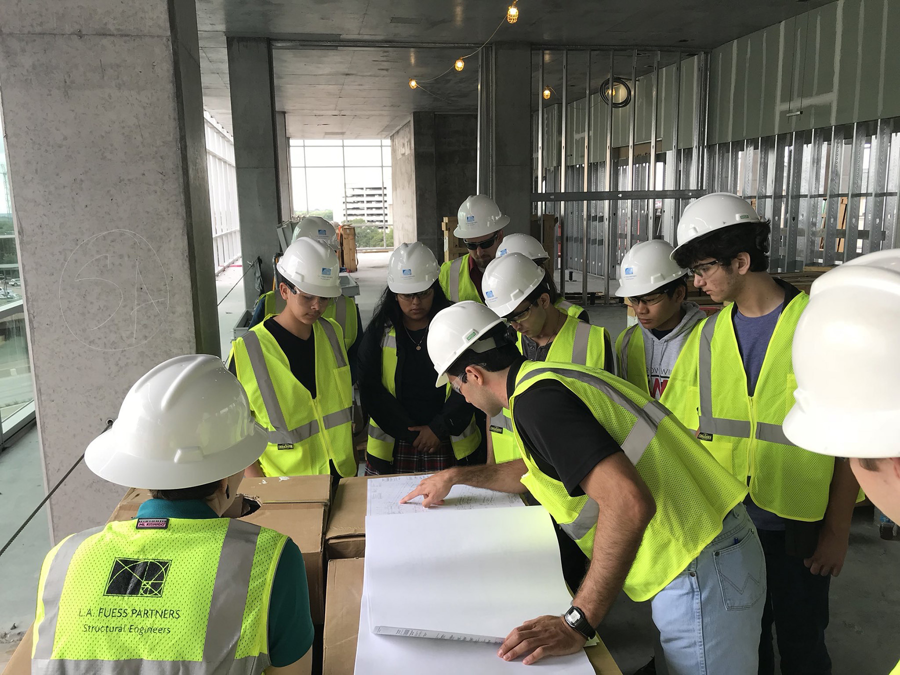 group of students and engineers wearing hard hats and safety vests looking at a drawing