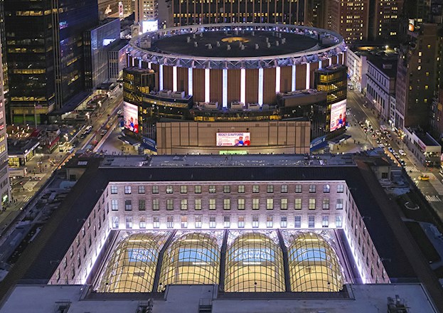 cityscape showing skyscrapers, midrise buildings, and low-rise buildings, a circular building that has signs on it, and a rectangular building with a sunken section that is topped by glass. 