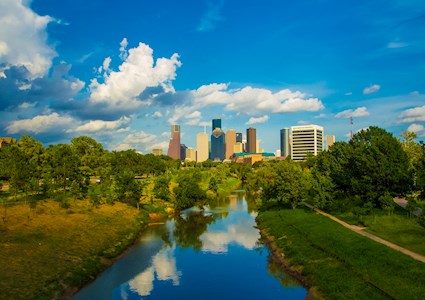 photo showing the Houston skyline in the background and a waterway surrounded by trees in the foreground lead 