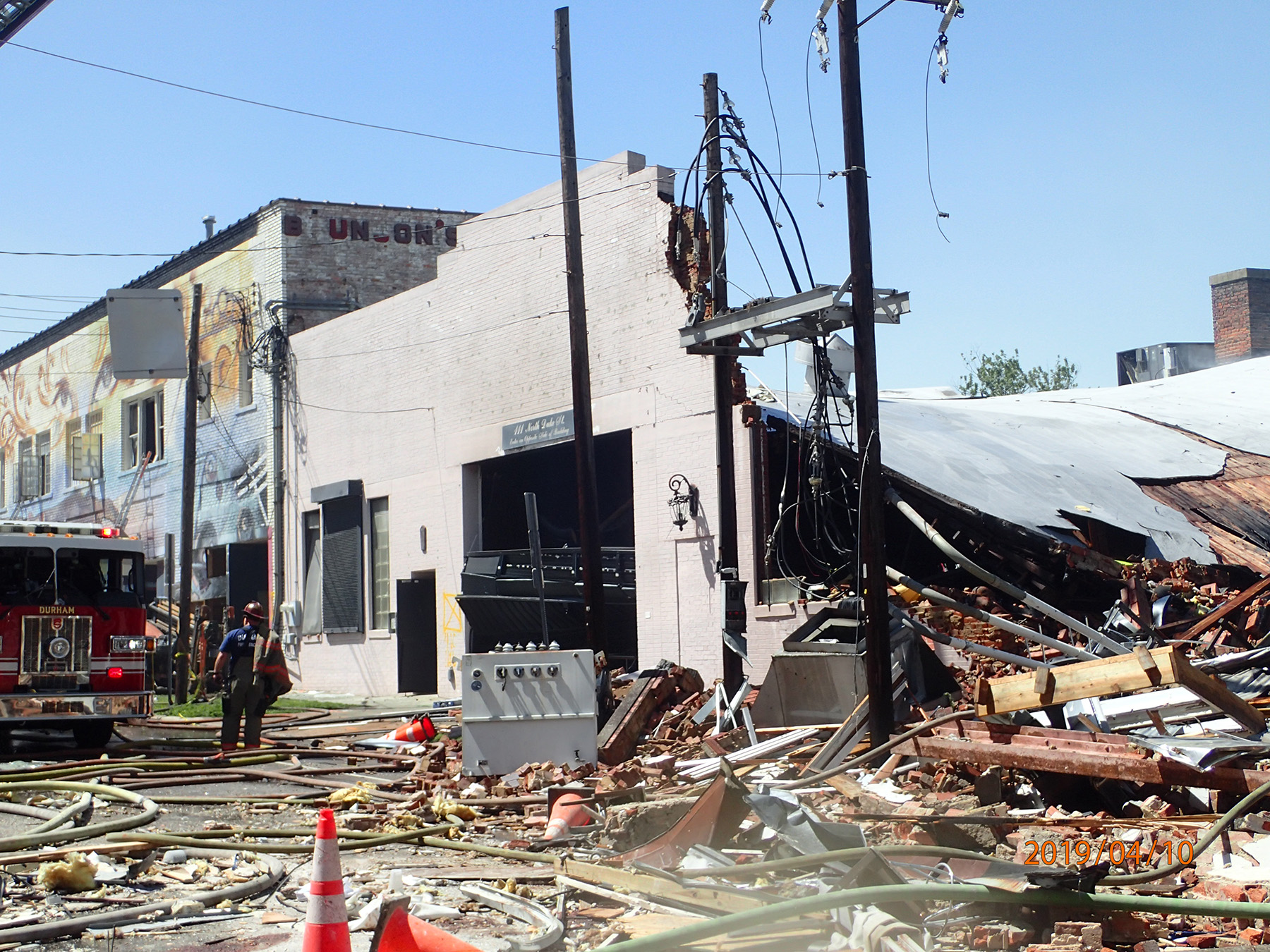 partially destroyed building with lots of debris, damaged utility poles. two firemen are seen in the background