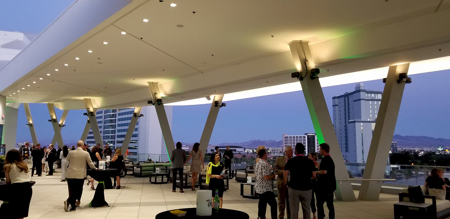 People standing around tables eating and drinking. In the background are V-shaped beams connected to the floor that support the roof