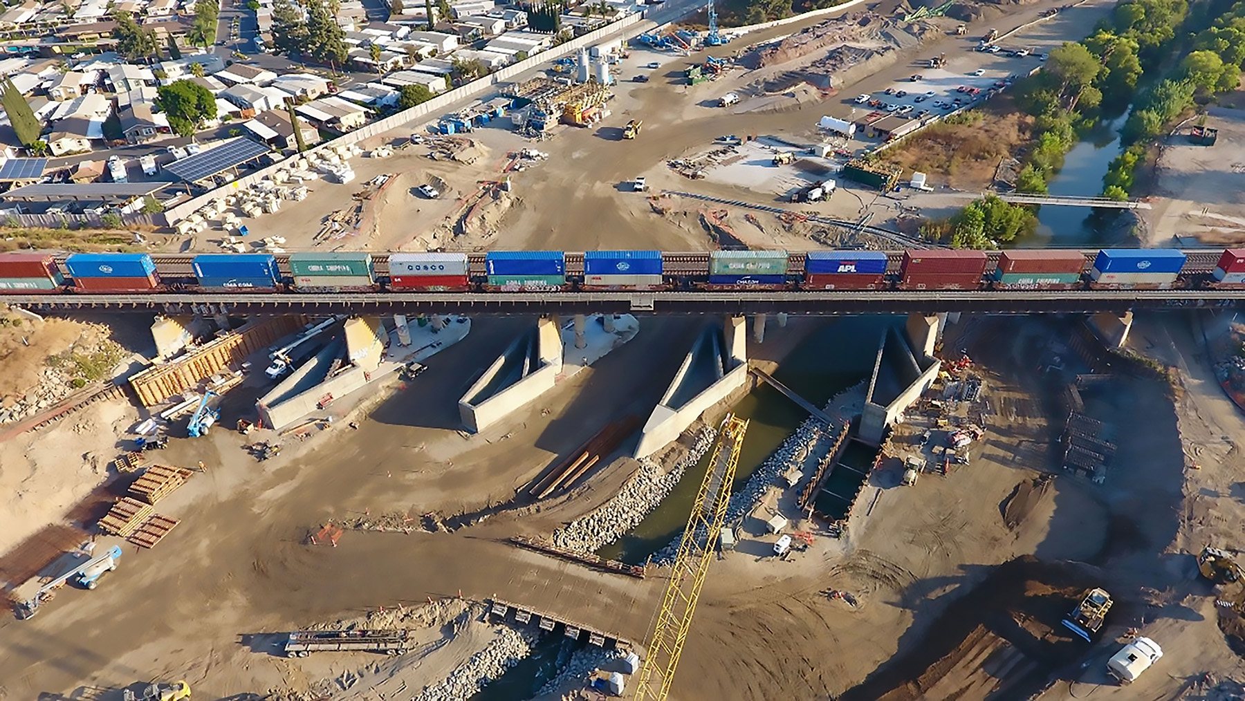 A train moves along a railway bridge. Underneath is a dry riverbed in which flood-prevention elements are being installed. 