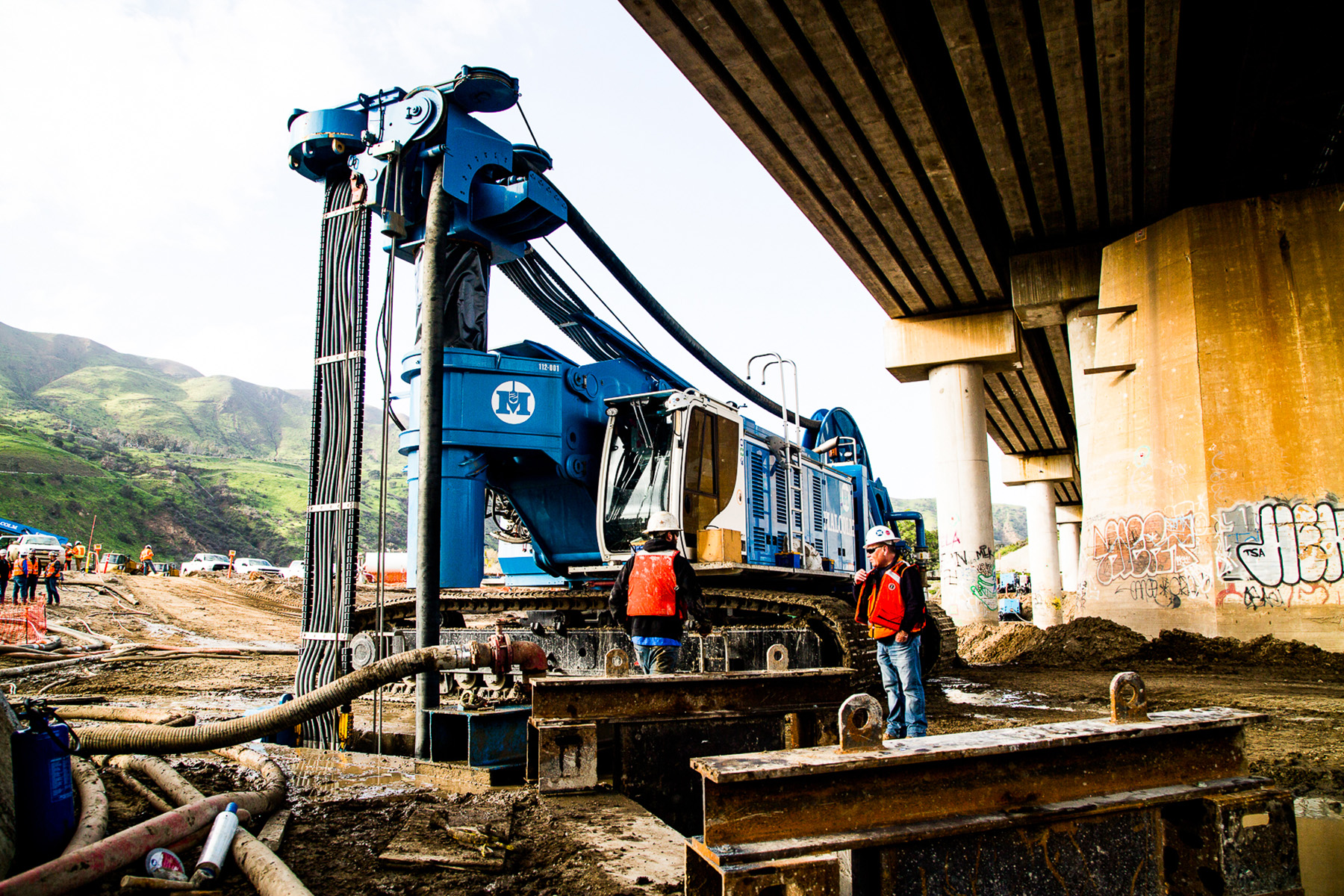 Workers stand next to a large blue machine placed under a bridge. 