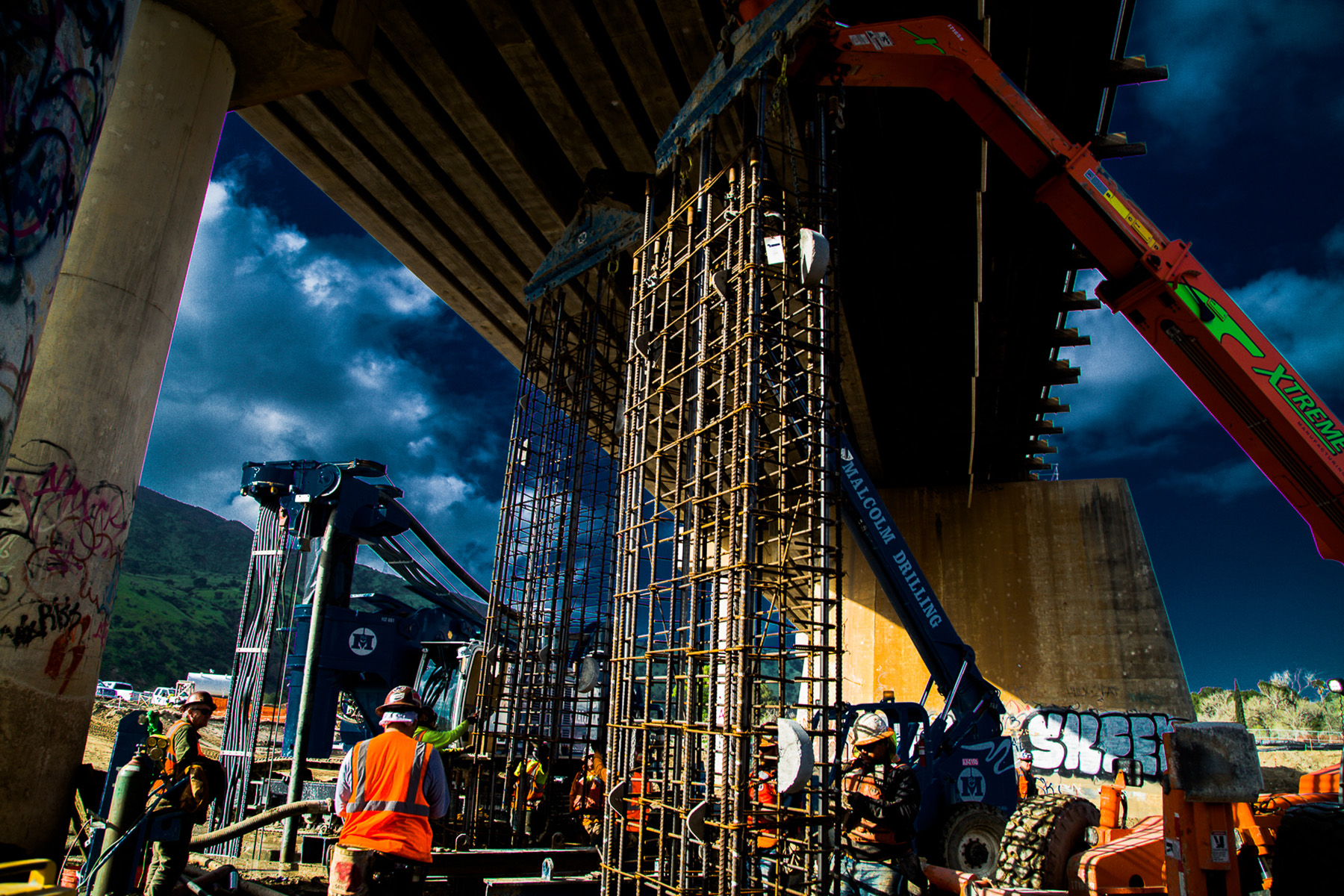 Workers install tall steel panels near the base of a bridge. 