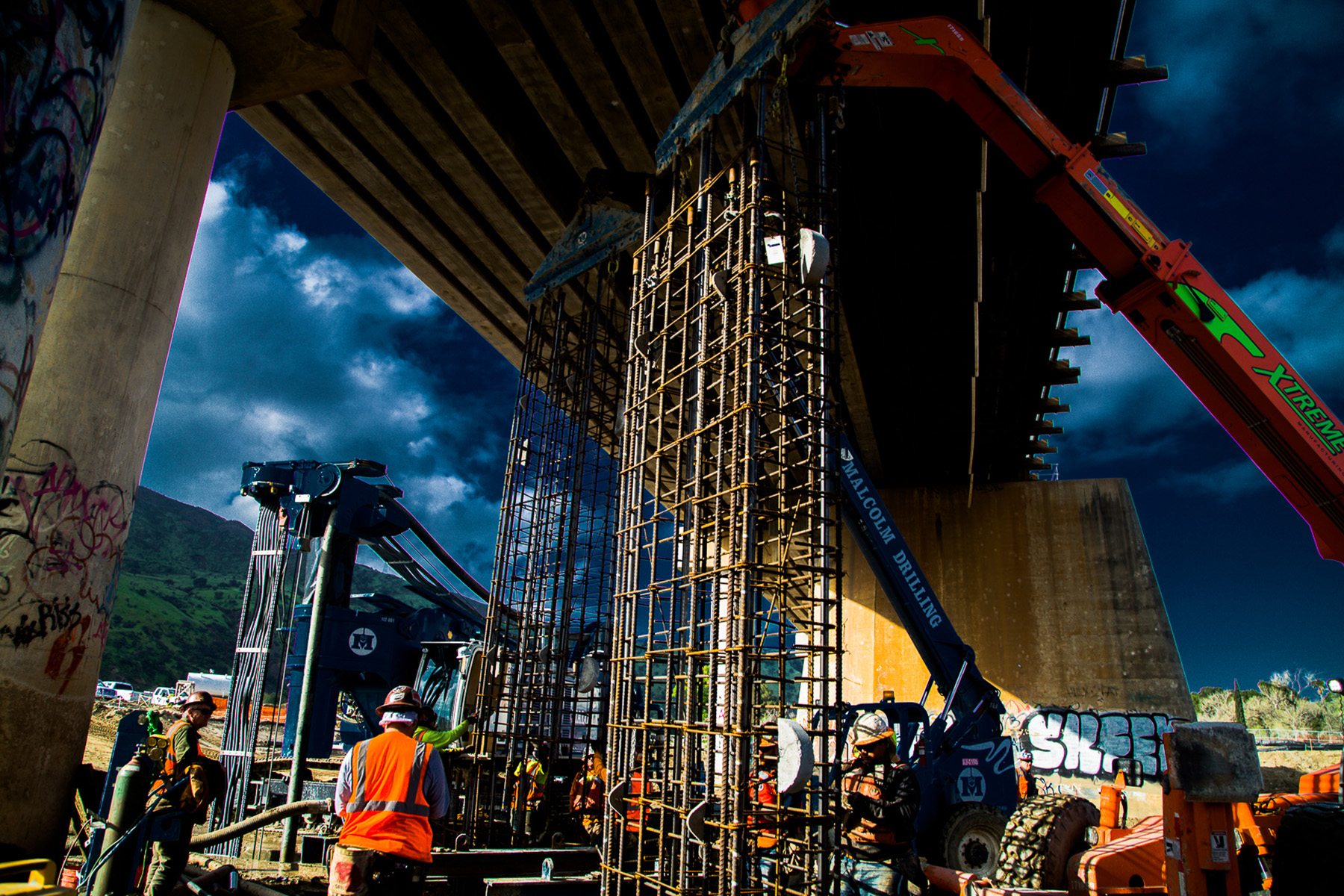 Workers install tall steel panels near the base of a bridge.