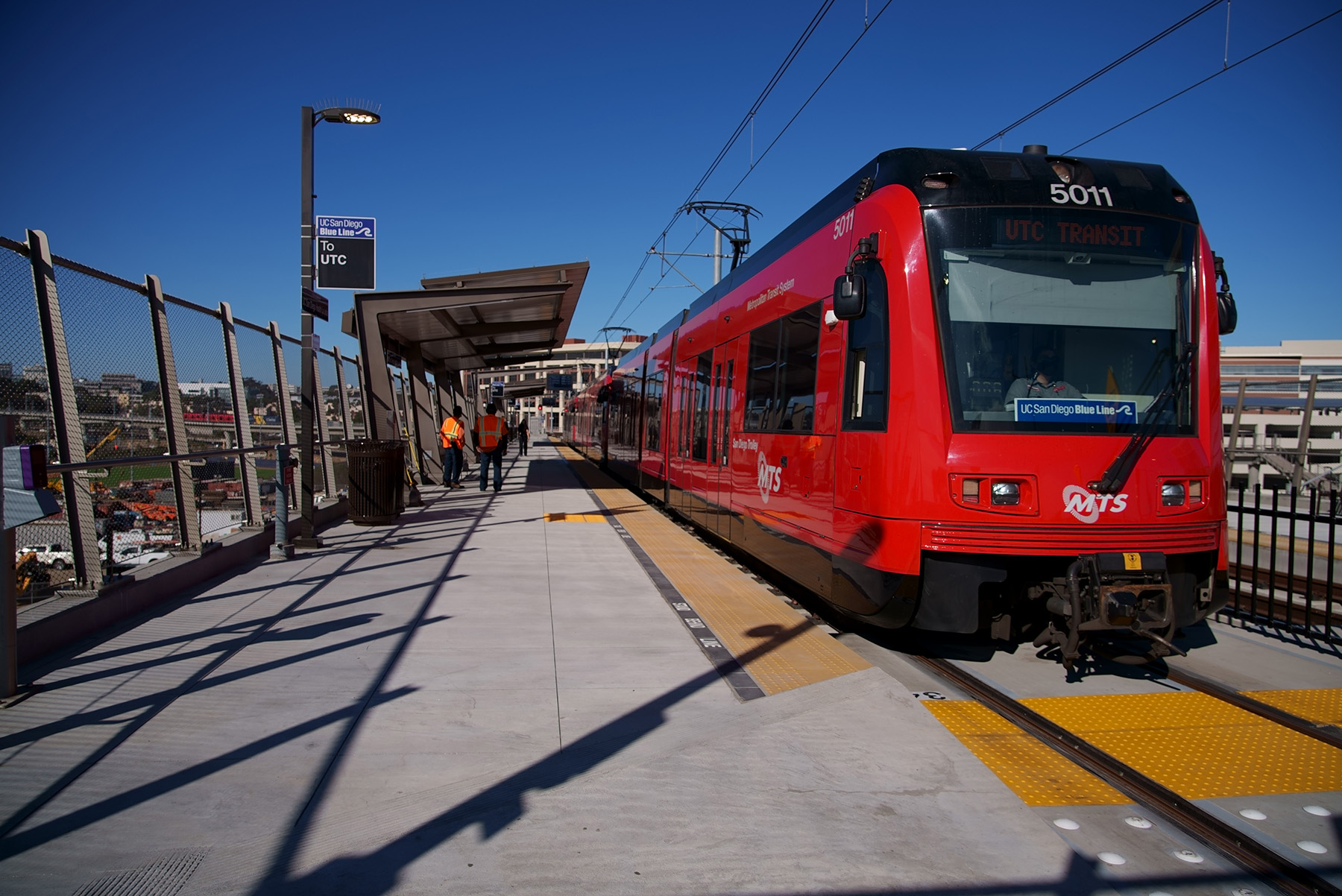 Train stopped at a trolley station. People in safety vests are on the platform.