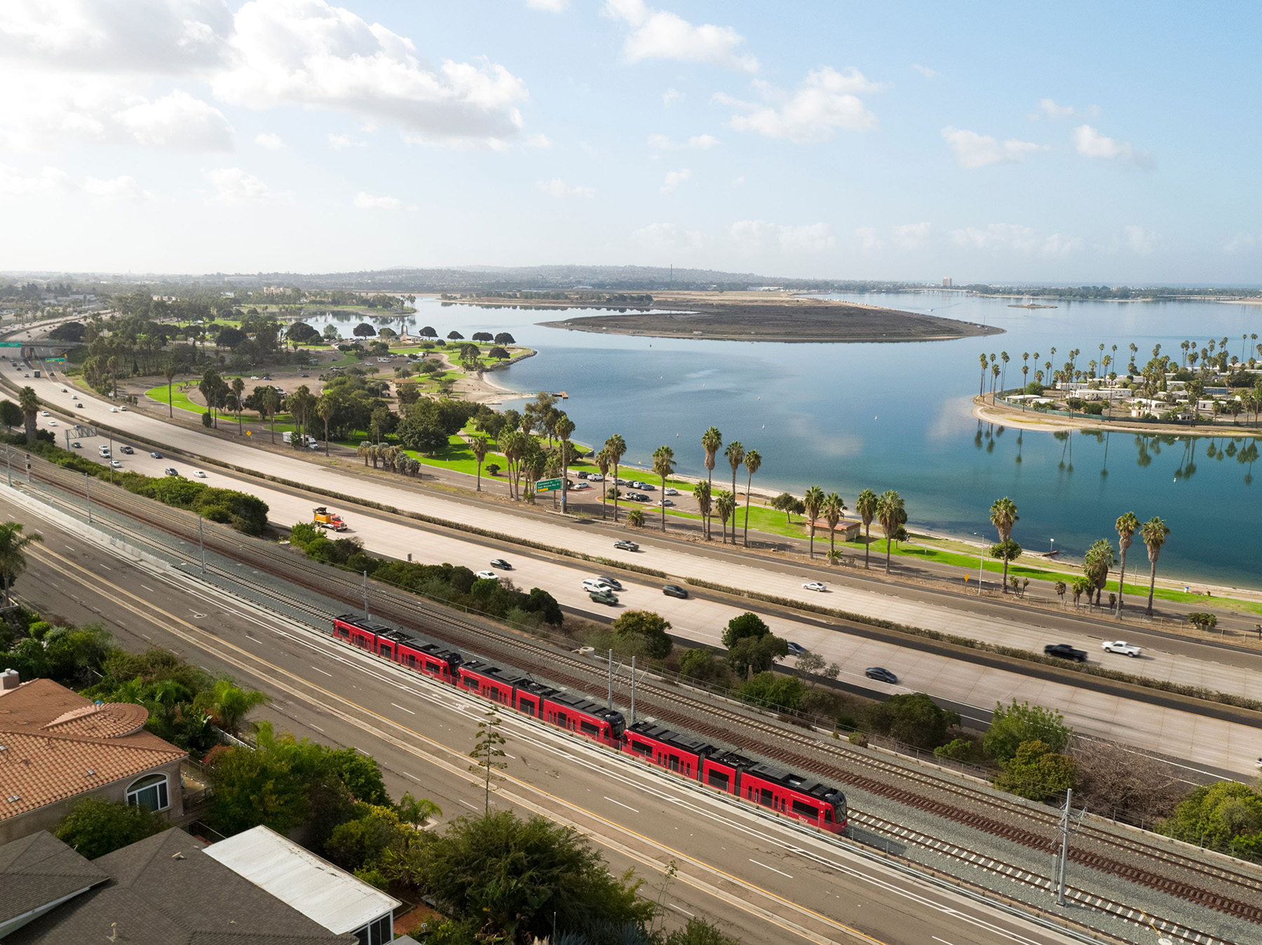 Multicar red train moves along the tracks. Water and palm trees are in the background.