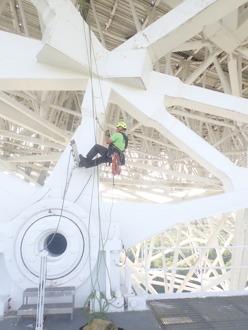 Man in green shirt and black pants is holding ropes as he inspects a part of a telescope.