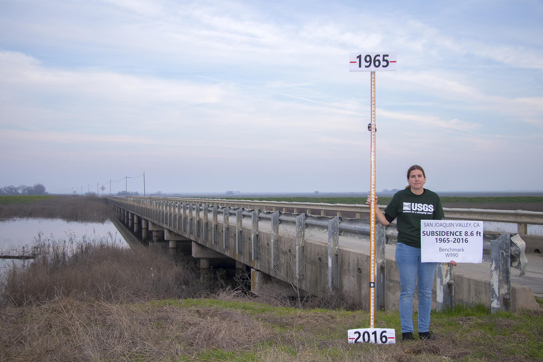 WOMAN IN USGS TOP SHOWING SUBSIDENCE LEVELS WITH MEASURING STICK