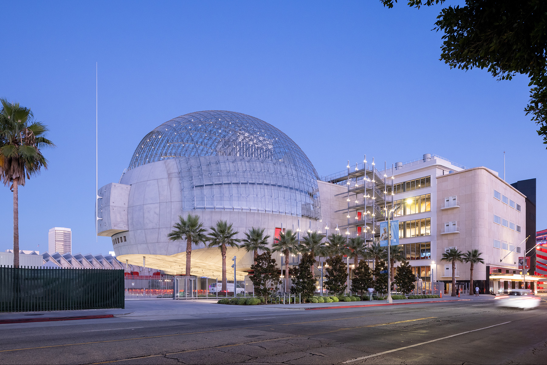 Large round structure with concrete base topped by a spherical glass roof. This building is connected by stairs to a multilevel building.