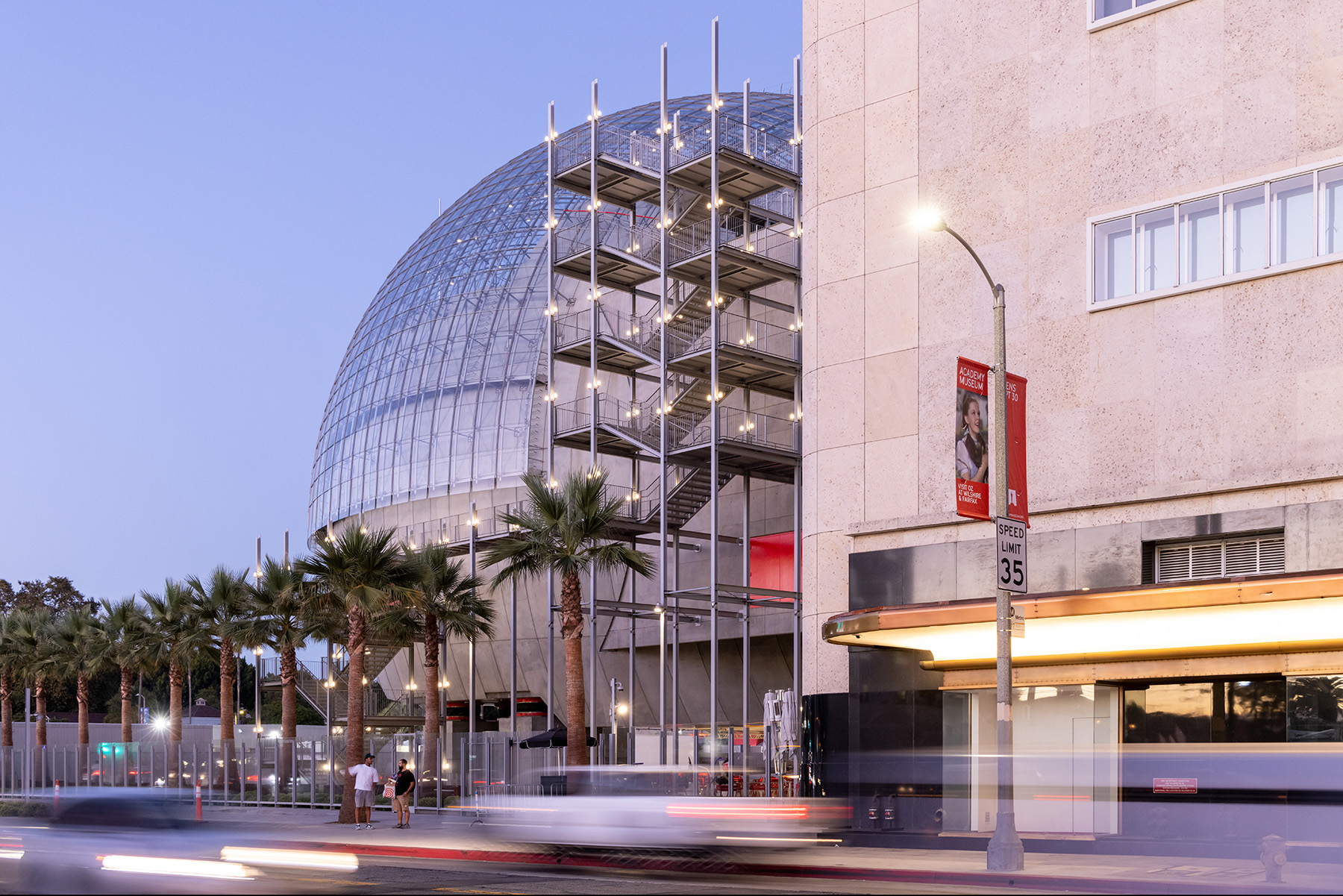 Large round structure with concrete base topped by a spherical glass roof. Stairs connect this building to another. There are palm trees in the foreground.