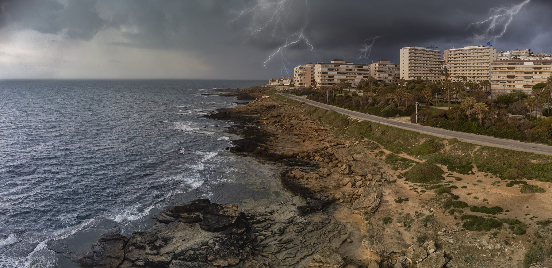 Ocean on the left, highway and buildings on the right depicting a coastal community. There is lightning in the background.