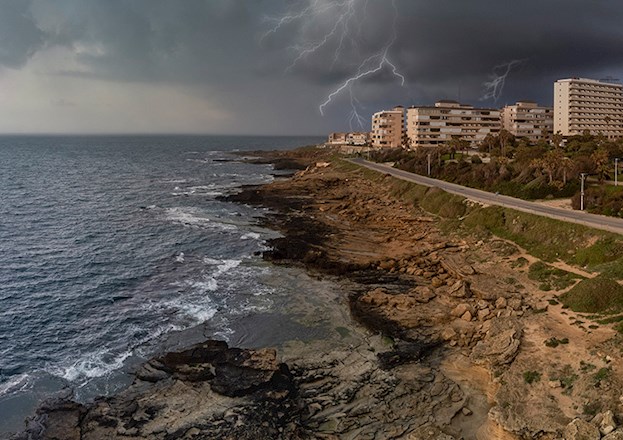 Ocean on the left, highway and buildings on the right depicting a coastal community. There is lightning in the background.