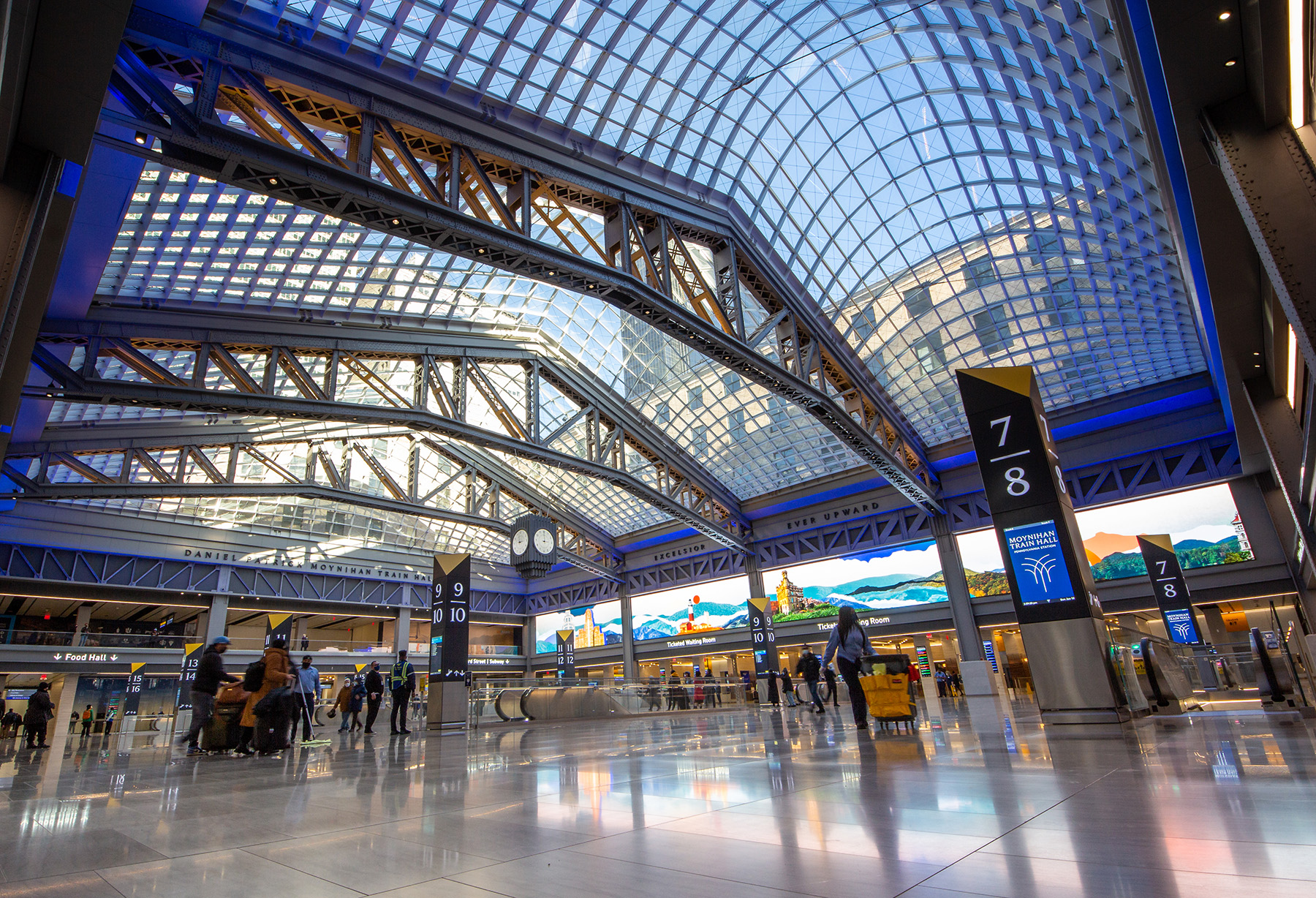 A new train hall is revealed showing the sky light supported by latticed steel trusses, open atrium, kiosks, and people with their luggage.