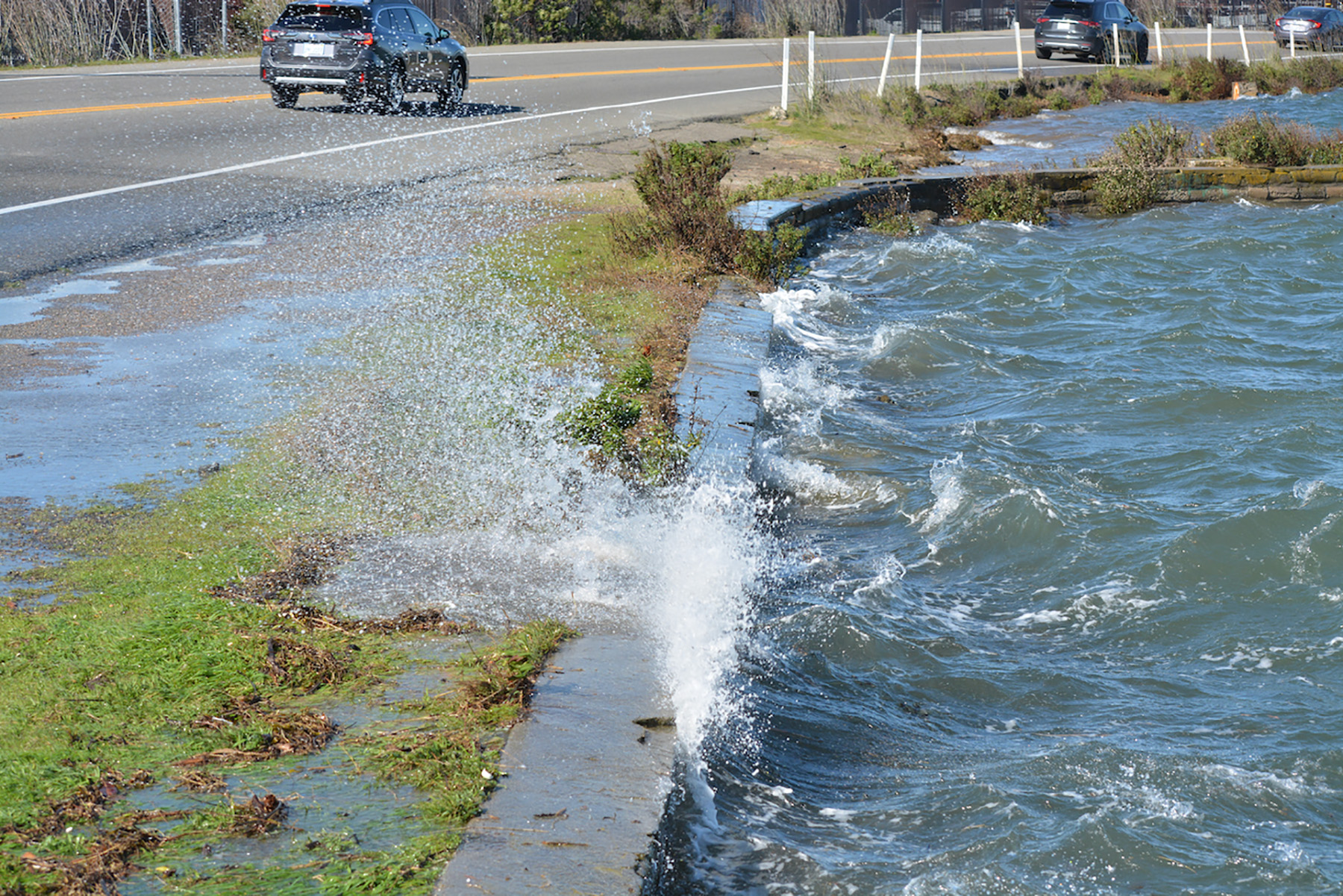 Photo shows water splashing against a rock and grass barrier that sits next to the roadway. 
