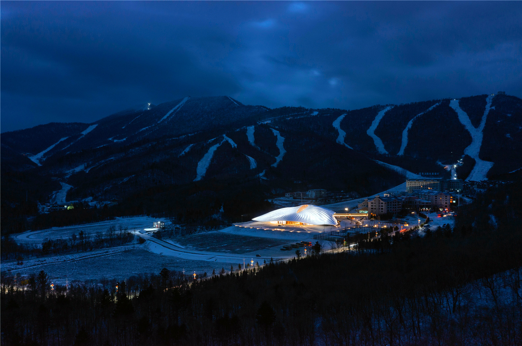 Nighttime image shows an irregular-shaped building lit up at night. The building is surrounded by mountains on one side and forest on the other. 