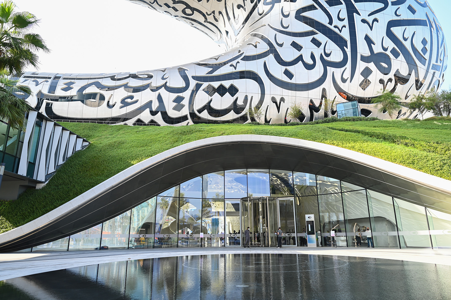 Entrance to a building is shown. You can also see a water feature in the foreground, walkway, and green mound that the building sits on.