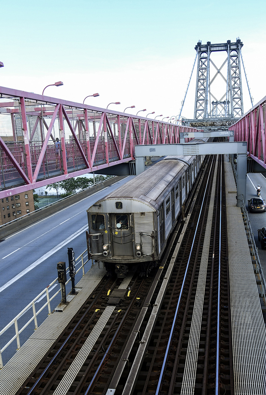 Image shows a train pulling out of a station. There is a pedestrian bridge to the left.