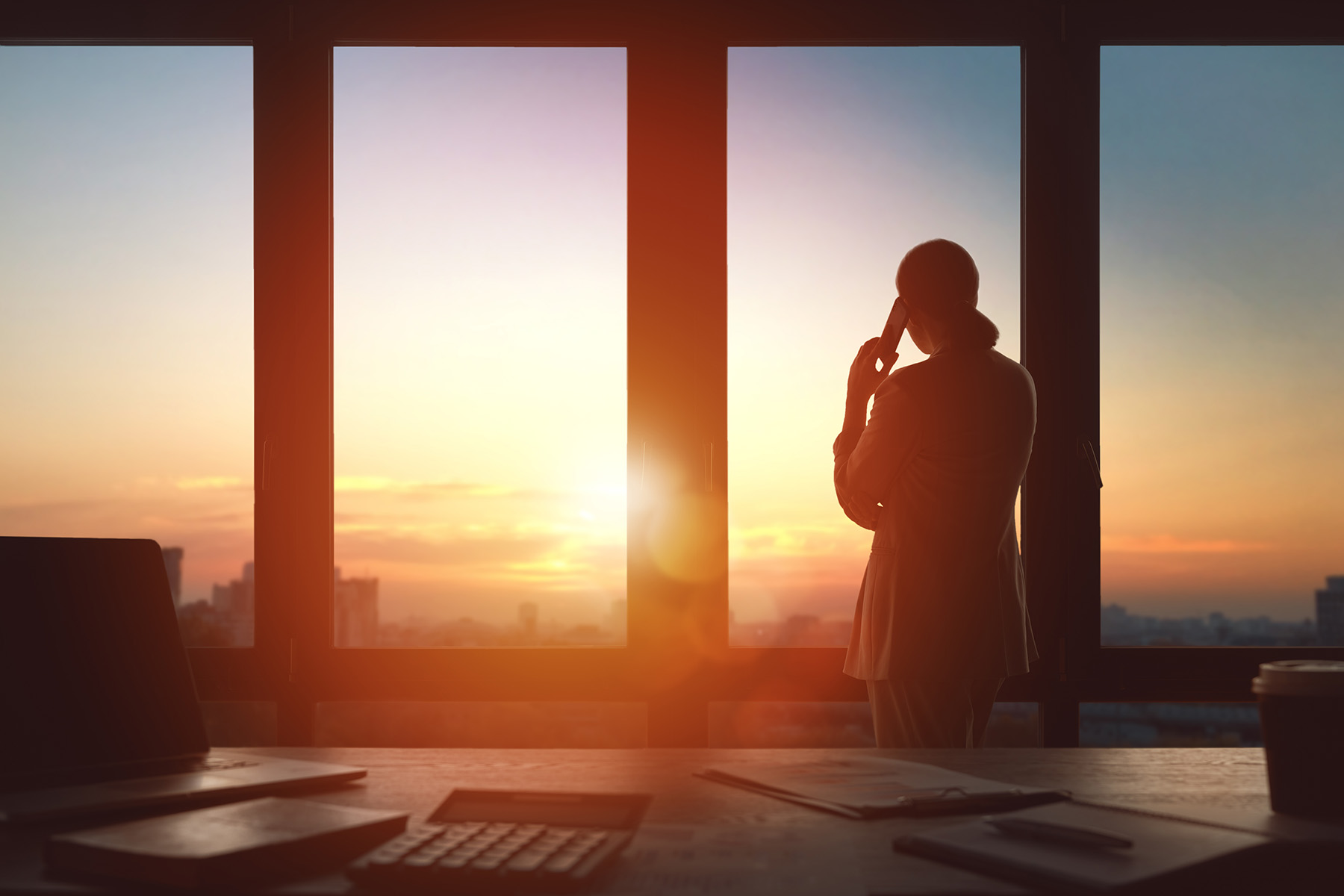 Woman stares out of a window as she talks on the phone. In the background is the outline of a city. 