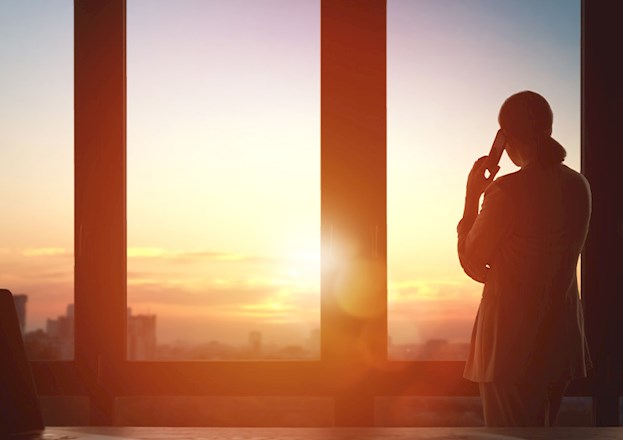 Woman stares out of a window as she talks on the phone. In the background is the outline of a city. 