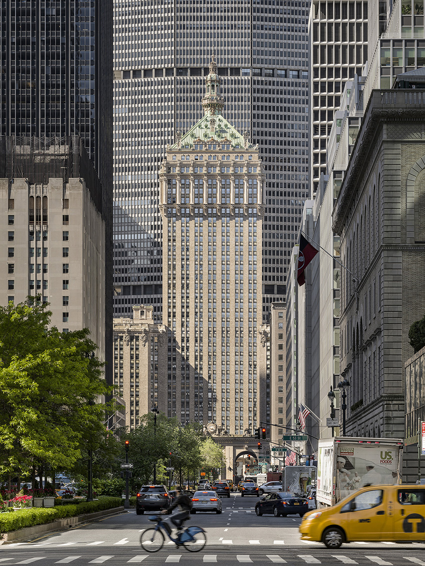 A high-rise building with a historic facade is shown. There are cars in the foreground and other tall buildings in the background.