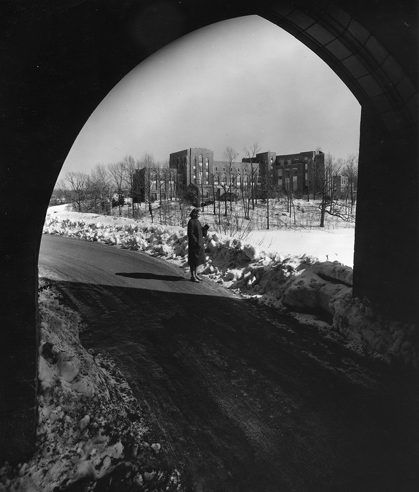 Black and white photos shows a woman in a coat standing on a road. There is snow on the side of the road. IN the background is a large building.