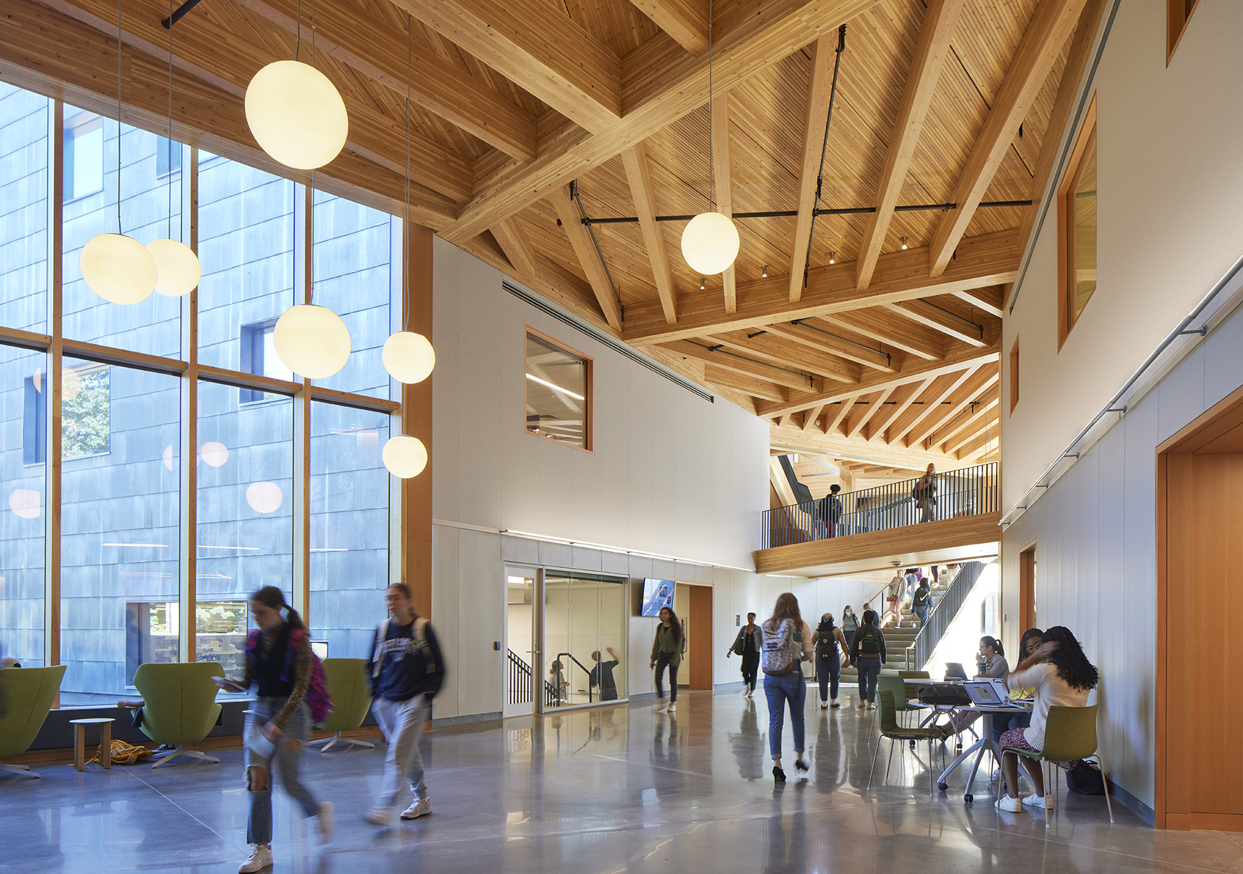 Students walk through a large multistory space. The ceiling is made of a light wood and the floor is concrete.