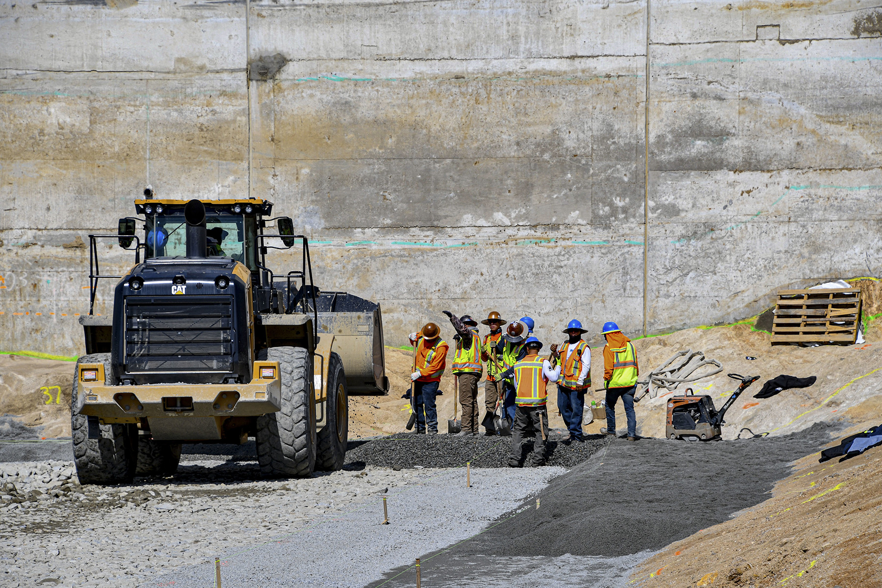 A vehicle with large wheels and a scooper in the front moves dirt around. Workers in hard hats and safety vests shovel the dirt.