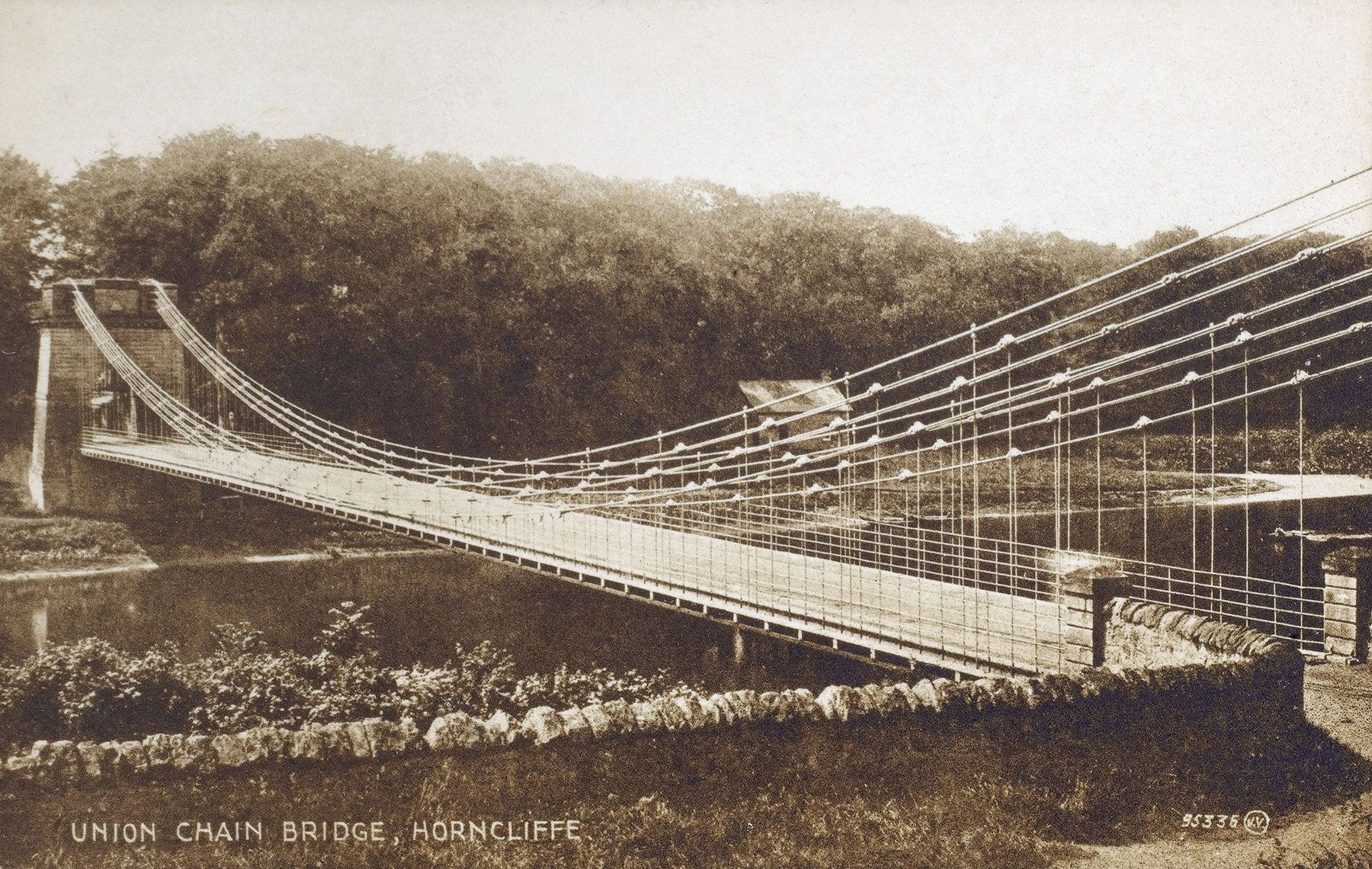 A sepia-toned picture shows the deck and piers of a bridge that spans a body of water. The suspension system comprises iron rods and chains. 