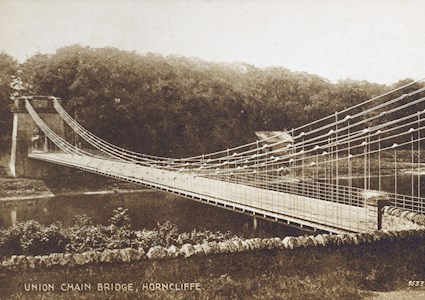 A sepia-toned picture shows the deck and piers of a bridge that spans a body of water. The suspension system comprises iron rods and chains. 