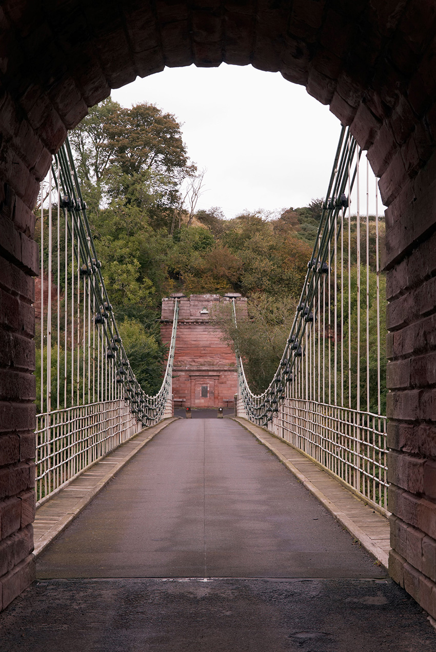 The photograph shows the deck of a bridge. On either side of the bridge deck are arced iron chains that support the deck and a series of iron rods. In the distance is a tall masonry pillar that the chains are connected to.