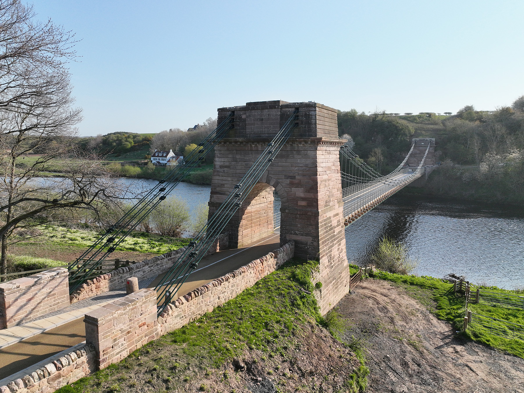 A tall masonry pier is shown in the foreground. Iron chains weave through the top of the pillar to attach to the ground on one side and a bridge suspension system on the other. In the background can be seen the other pillar the chains attach to. 