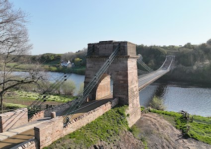 A tall masonry pier is shown in the foreground. Iron chains weave through the top of the pillar to attach to the ground on one side and a bridge suspension system on the other. In the background can be seen the other pillar the chains attach to.