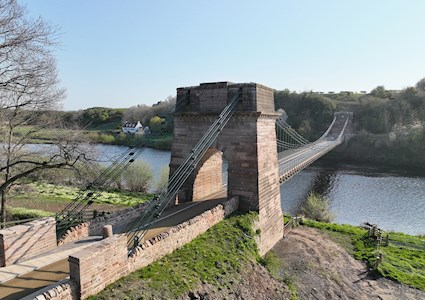 A tall masonry pier is shown in the foreground. Iron chains weave through the top of the pillar to attach to the ground on one side and a bridge suspension system on the other. In the background can be seen the other pillar the chains attach to.