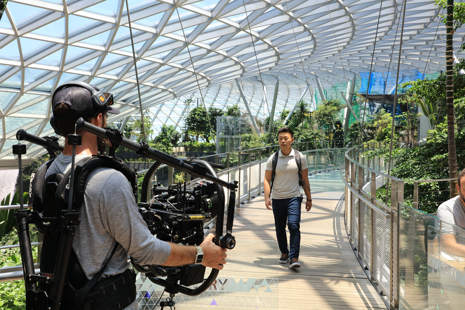 A man wearing jeans, t-shirt, and a backpack in an indoor atrium walks toward another man holding filming equipment. 